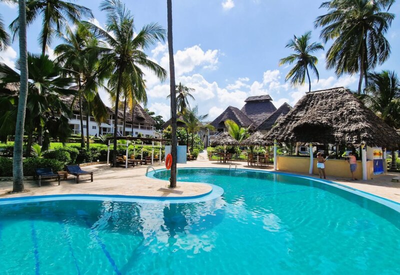 Resort pool surrounded by palm trees, thatched-roof bar, outdoor seating, and bungalow-style accommodations under a sunny blue sky
