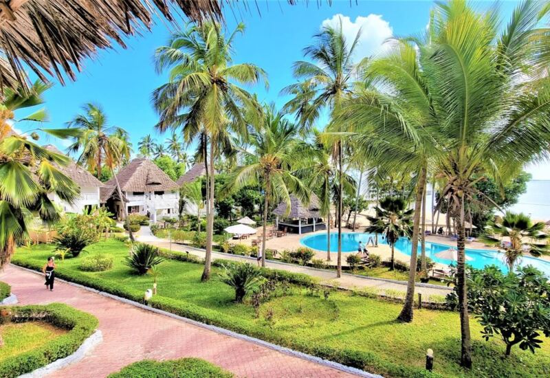 Resort with thatched-roof bungalows, palm trees, lush gardens, a round pool, and beach views under a bright blue sky