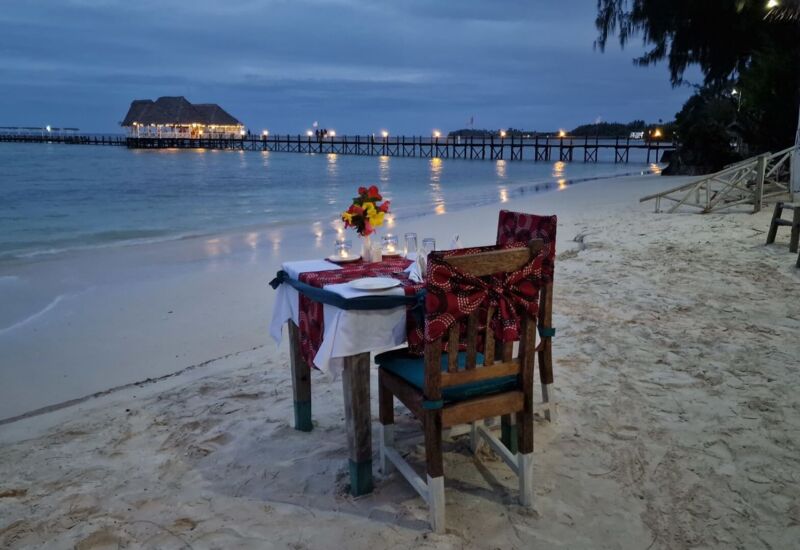 Romantic candlelit dinner table for two on a sandy beach at twilight with ocean view and distant pier restaurant lights
