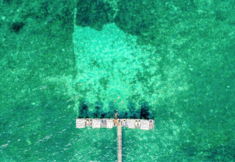 Aerial view of a wooden pier extending into clear turquoise ocean waters with sun loungers and shaded cabanas for relaxation