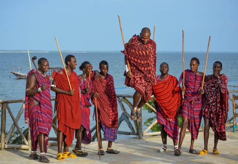 Group of Maasai men in traditional red and purple shukas perform a jumping dance by a lake under clear blue sky