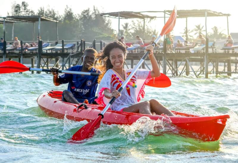 Couple enjoying kayaking in bright red tandem kayak on clear ocean waters near a pier with sunbathers on lounge chairs