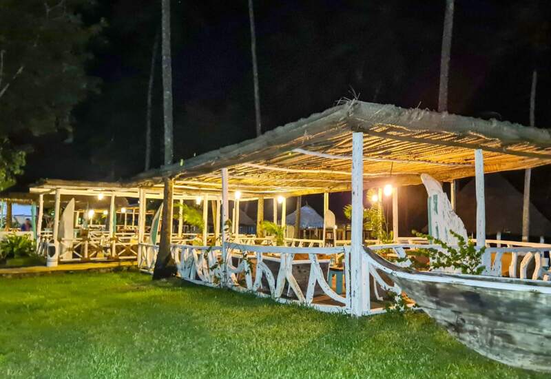 Outdoor tropical dining area at night with wooden pergolas, warm lighting, palm trees, and rustic boat decor on lush green lawn
