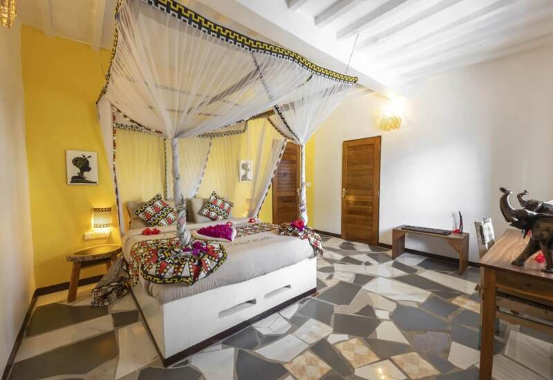 Bright room with a four-poster bed draped in white mosquito netting, vibrant African-patterned pillows, and decorative touches on a polished tile floor