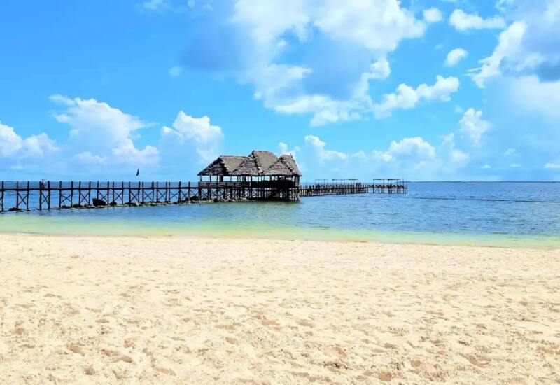 Sandy beach with clear turquoise water leading to a wooden pier and thatched-roof pavilion over the ocean under a bright blue sky