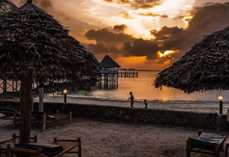 Beachfront resort at sunset with thatched umbrellas, lounge chairs, calm water, and children playing along the shore