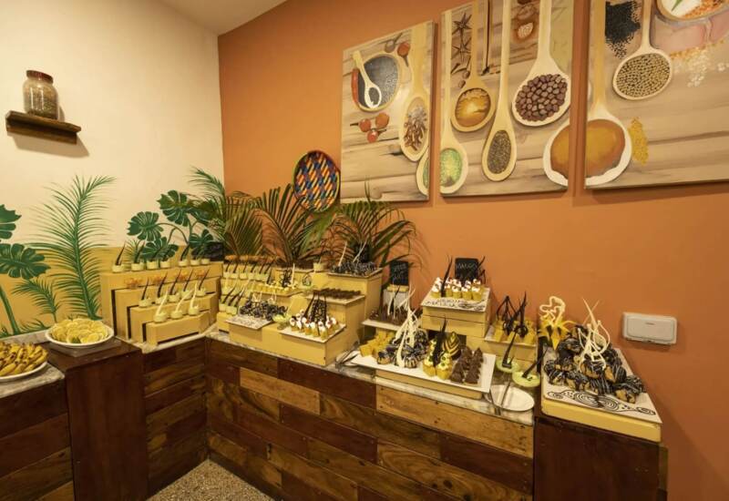 Rustic dessert buffet with assorted pastries and sweets displayed on wooden counters against a warm orange wall with tropical plant decor
