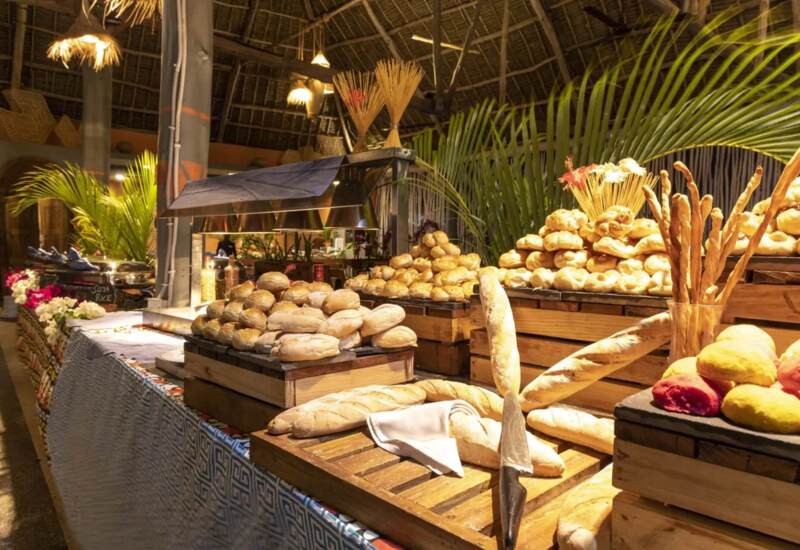 Rustic buffet table featuring a variety of fresh breads and rolls under a thatched roof, surrounded by tropical plants
