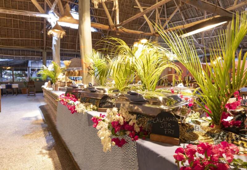 Buffet setup under a rustic thatched roof with vibrant flowers, palm leaves, and covered chafing dishes labeled with menu items
