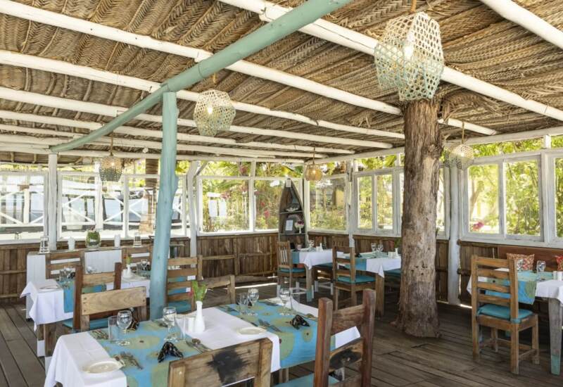 Rustic open-air restaurant with wooden furniture, white tablecloths, turquoise accents, woven ceiling, and natural tree trunk feature