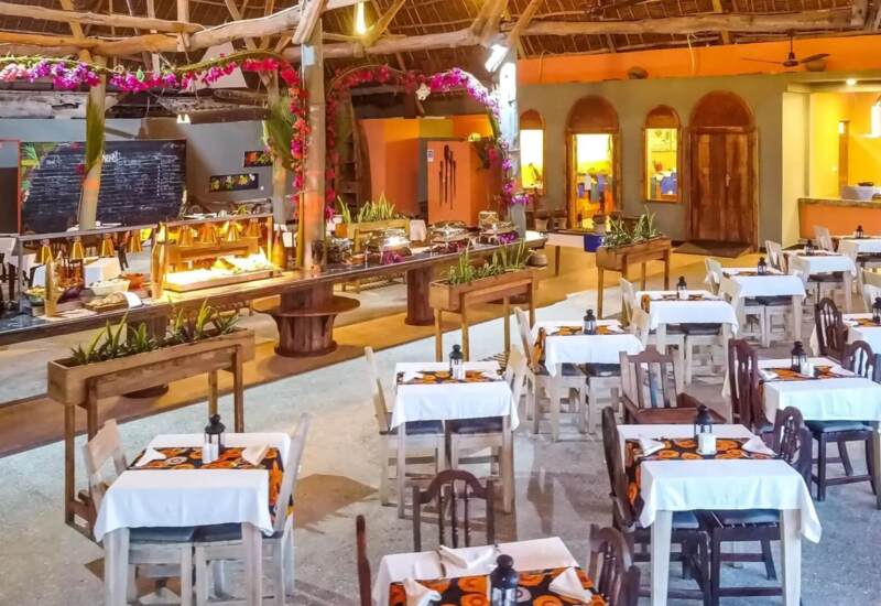 Open-air hotel restaurant with white linen-covered tables, wooden chairs, floral buffet arches, and a bright, rustic ambiance