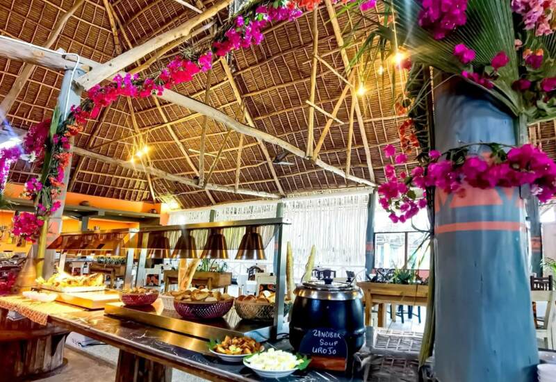 Open-air buffet under a thatched roof with local bread, soup station, tropical flowers, and warm ambient lighting