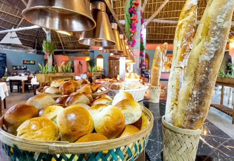 Freshly baked bread rolls and baguettes displayed under warm lights in a rustic hotel restaurant with thatched ceiling and cozy seating