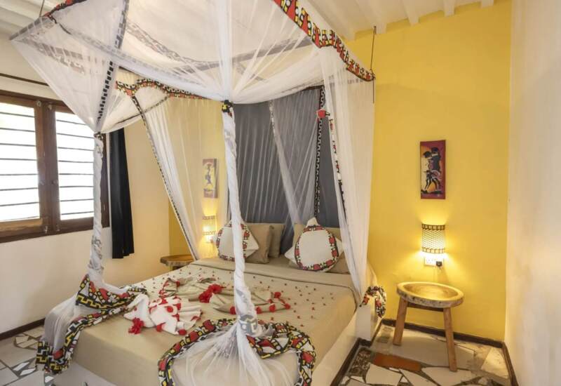 Cozy hotel room with a canopy bed draped in white mosquito netting, yellow walls, wooden accents, and decorative pillows.