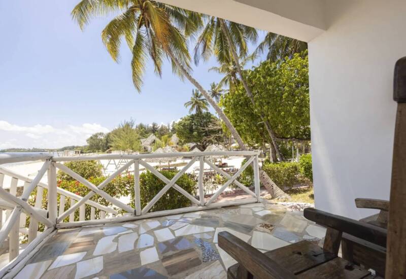 Beachfront patio with wooden chairs, mosaic tile floor, white wooden railing, palm trees, and sandy shore views