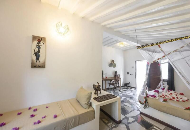 Bright hotel room with white beamed ceiling, mosaic tile floor, daybed, decorative elephant sculptures, and bed with mosquito net and floral accents