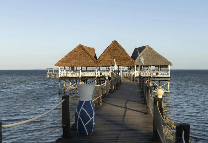 Wooden pier leading to a stilted overwater restaurant with thatched roofs, overlooking calm ocean waters under a clear sky