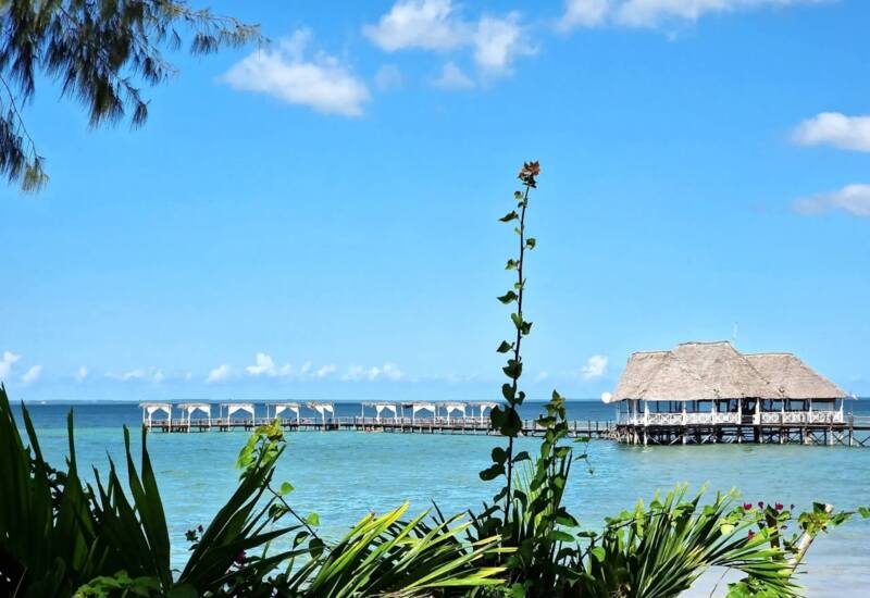 Thatched-roof overwater pavilion connected by a wooden pier lined with shaded cabanas on a clear tropical ocean under blue sky