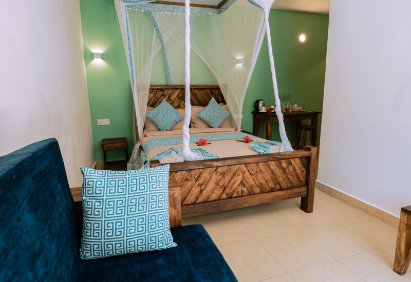 Cozy hotel room with a wooden bed featuring patterned pillows, a mosquito net, a blue sofa, and a small table with water bottles and a kettle