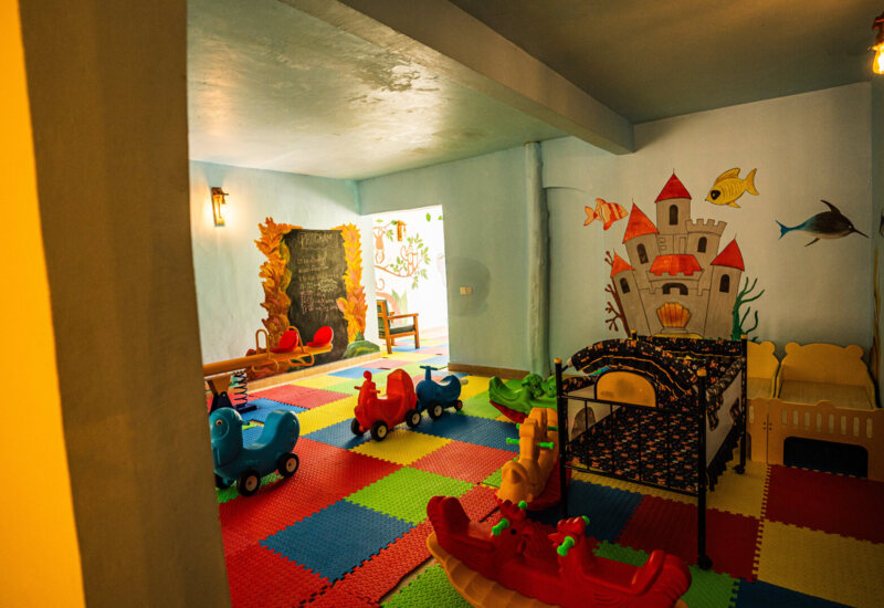 Colorful indoor kids' playroom with foam floor mats, small ride-on toys, wall murals of castle and sea creatures, and child-sized furniture