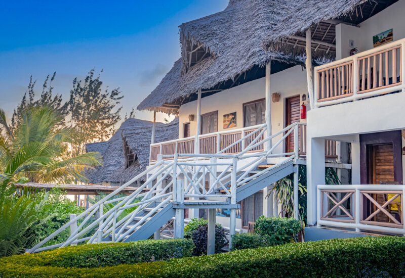 Tropical hotel with thatched roofs, wooden balconies, lush greenery, and stairs leading to upper guest rooms at sunset