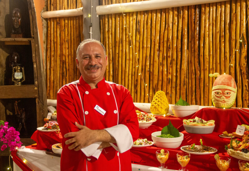 Smiling chef in red uniform stands beside a festive buffet with carved fruit and assorted dishes on red-draped tables