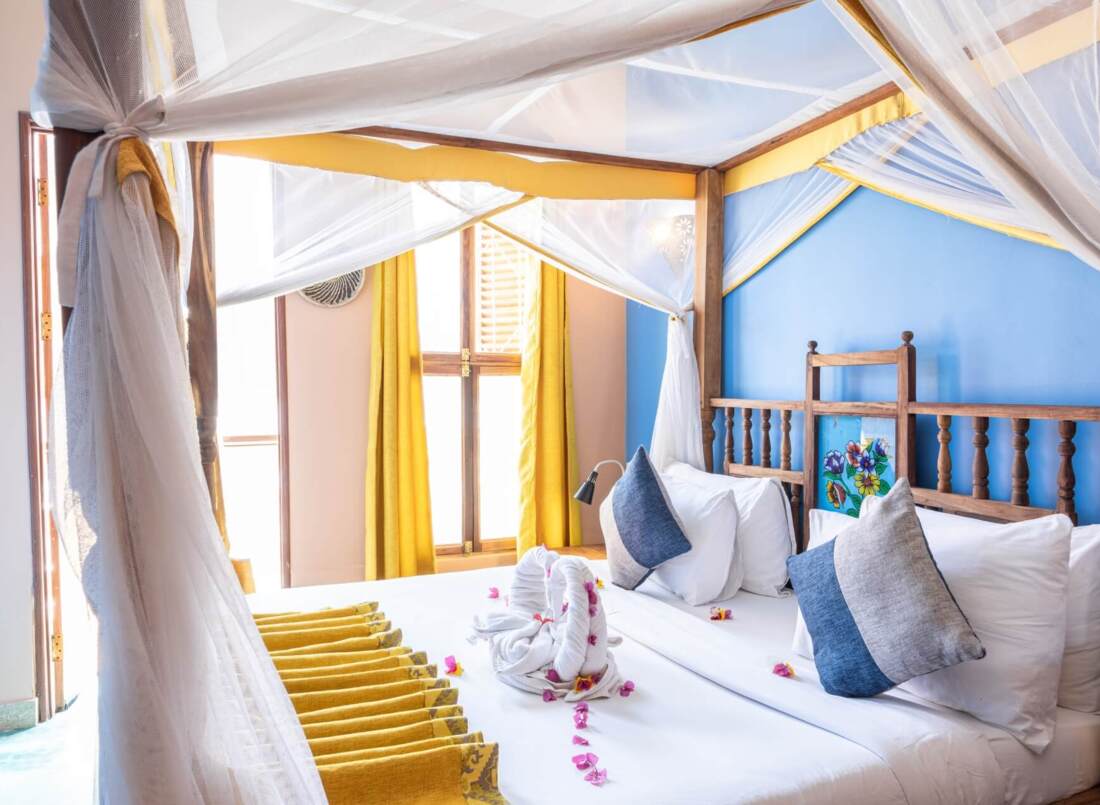 Bright four-poster bed with white linens, blue and gray pillows, yellow curtains, and towel swans with flower petals in airy room