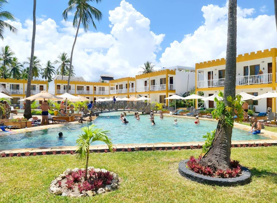 Tropical resort with yellow-accented rooms surrounding a large pool where guests enjoy swimming and lounging under umbrellas