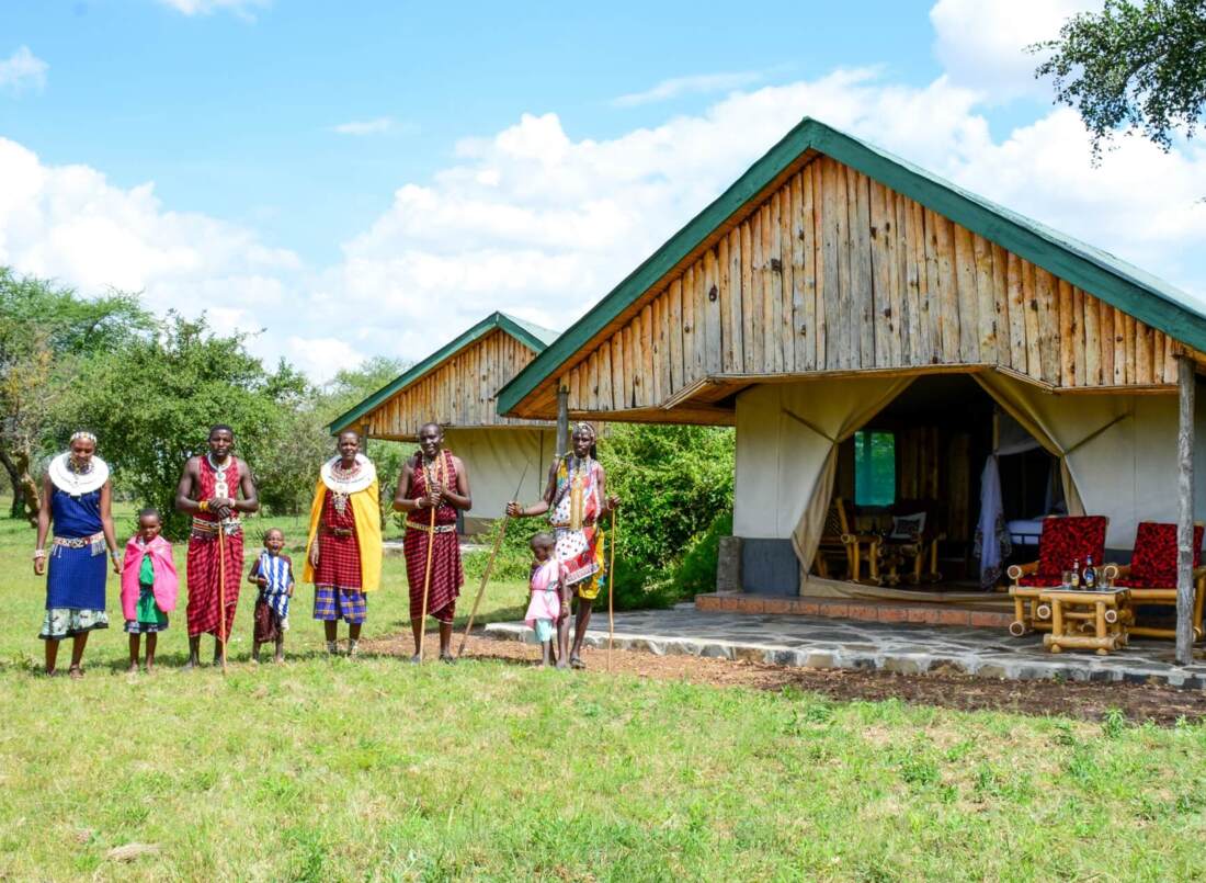 Rustic safari tents with wooden roofs and outdoor seating in a grassy area, with a group of people in traditional Maasai attire nearby