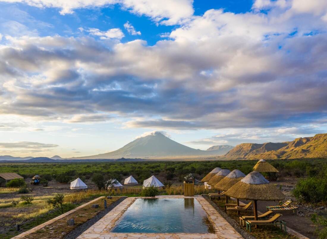 Outdoor pool with stone deck and thatched umbrellas overlooking safari tents and a mountain under a partly cloudy sky
