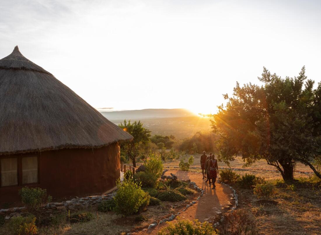 Traditional thatched-roof hut in a natural setting with guests walking on a stone-lined path at sunset