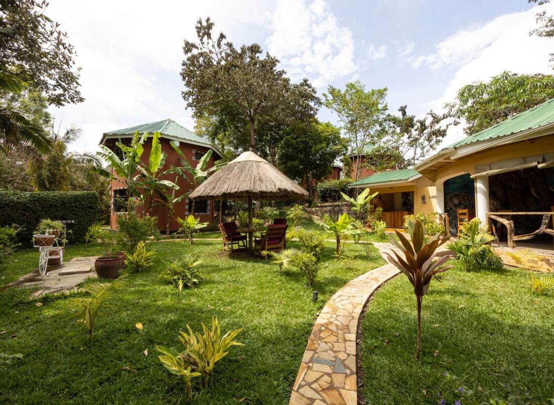 Tropical hotel garden with lush greenery, thatched umbrella seating area, stone pathway, and cottages with green metal roofs