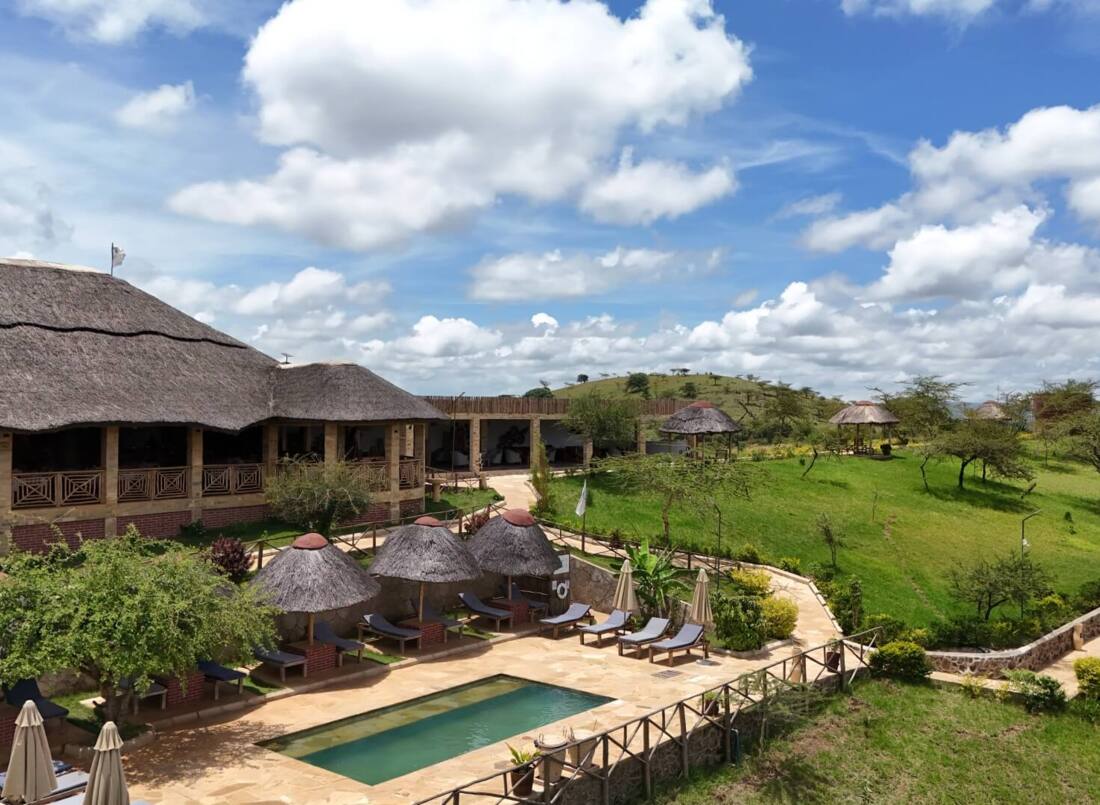 Thatched-roof lodge with outdoor seating and small pool, surrounded by green hills and clear blue sky with scattered clouds