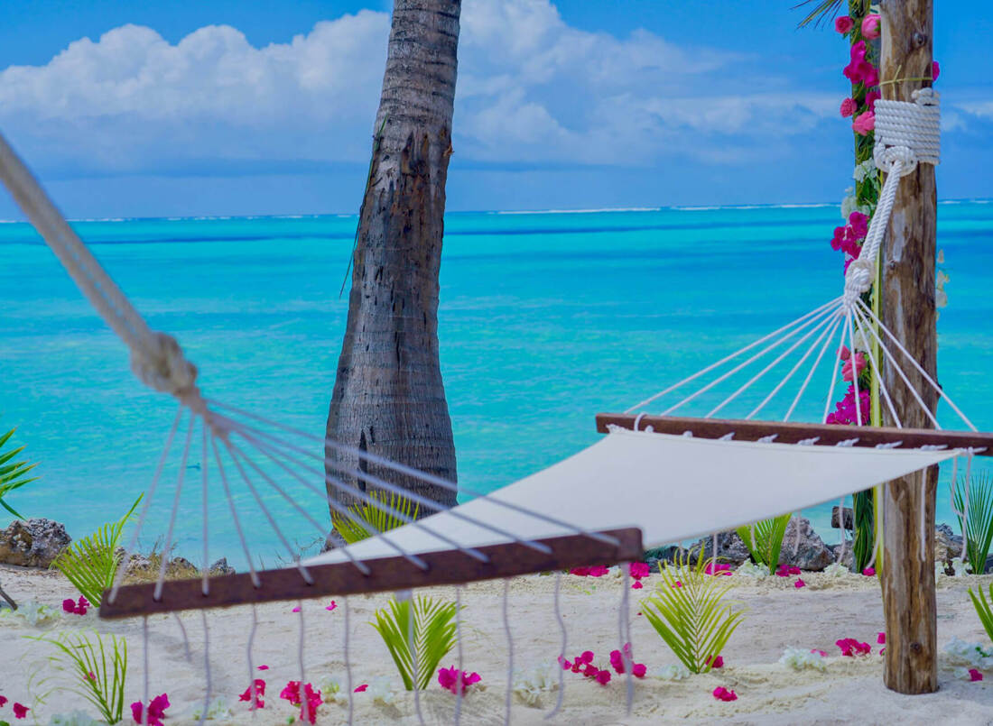 White hammock tied between wooden posts on a sandy beach with tropical flowers, palm trees, and turquoise ocean view