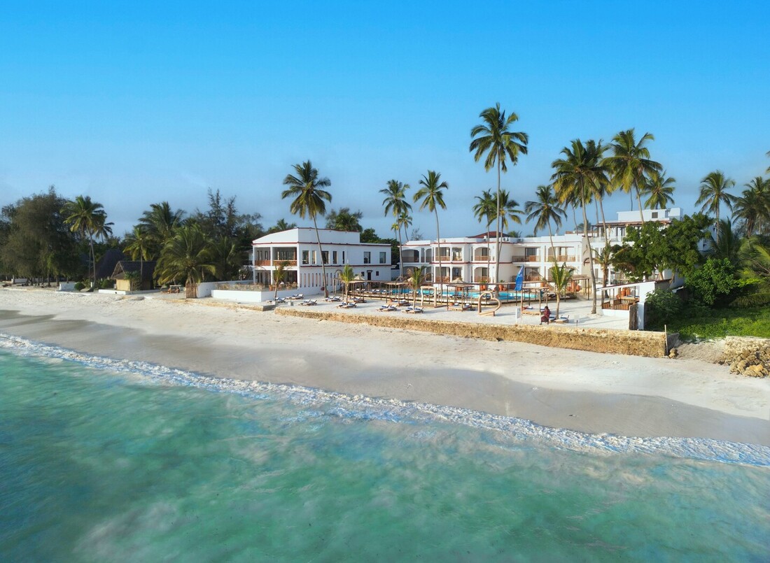 Oceanfront resort with white two-story buildings and palm trees, sandy beach, turquoise sea, poolside loungers and shaded cabanas.