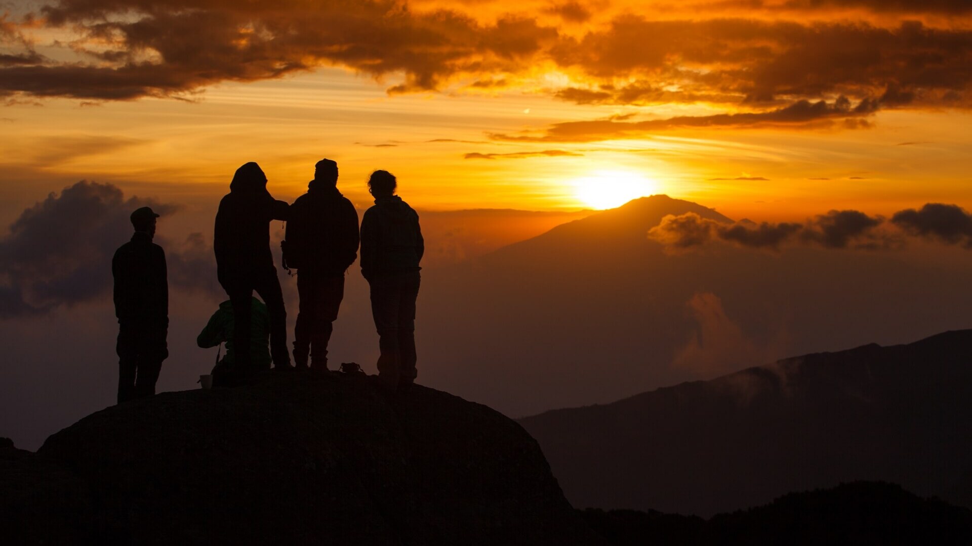 Group of hikers silhouetted on a mountain peak enjoying a dramatic orange sunset with distant clouds and mountain views