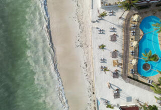Aerial view of a beachfront hotel with turquoise pool, palm trees, shaded cabanas, and lounge chairs on white sand by the ocean