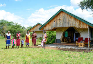 Eco-friendly safari tents with wooden roofs and outdoor seating, surrounded by lush greenery and a group of traditionally dressed Maasai people