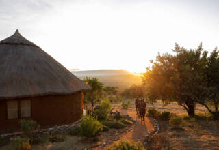 Sunset view of a traditional thatched round hut with a stone-bordered path leading through lush greenery and guests walking outdoors