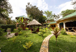 Tropical garden with lush greenery, winding stone path, thatched umbrella with seating, and guest cottages with green metal roofs