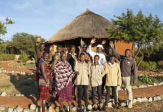 Group of hospitality staff smiling and waving outside a traditional thatched-roof lodge surrounded by greenery and pathways