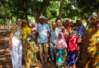 Smiling guests and local family in colorful traditional dress under shady trees, wearing shell garlands during a village welcome.