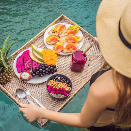 Woman in sunhat holding tray with tropical fruit, smoothie, granola bowl, and bruschetta by the pool for a relaxing breakfast