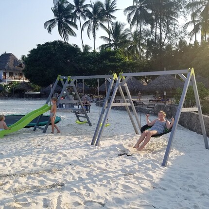 Beachfront resort playground on white sand with swings, slide and trampoline; children, palm trees and thatched bungalows.