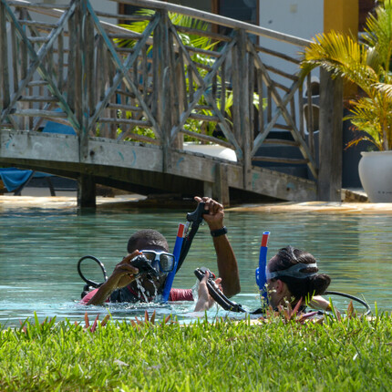 Resort pool scuba lesson with instructor and guest practicing masks and snorkels beside a wooden bridge and tropical plants