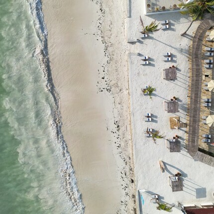 Aerial beachfront resort: curved turquoise pool with palm islets, white sand beach with palapas, sunbeds and waves