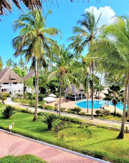 Tropical beachfront resort with thatched-roof villas, lush palm gardens, a circular pool, sun loungers, and guests enjoying the sunny pathways