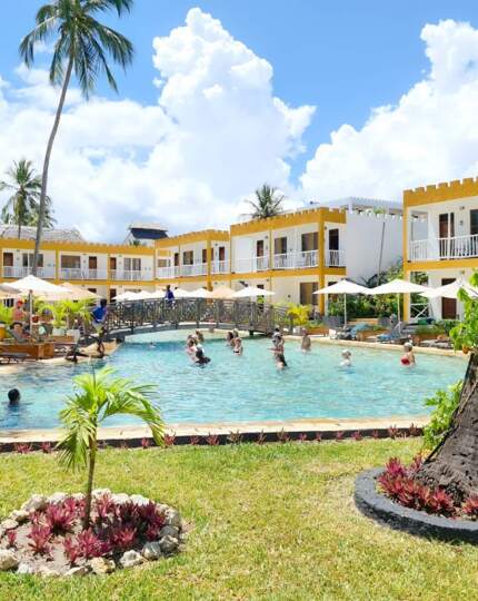 Tropical resort with yellow-accented rooms surrounding a large pool where guests enjoy swimming and lounging under umbrellas