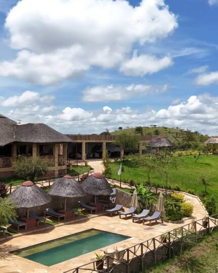 Thatched-roof lodge with outdoor seating and small pool, surrounded by green hills and clear blue sky with scattered clouds