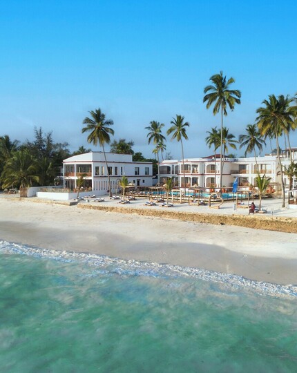 Oceanfront resort with white two-story buildings and palm trees, sandy beach, turquoise sea, poolside loungers and shaded cabanas.